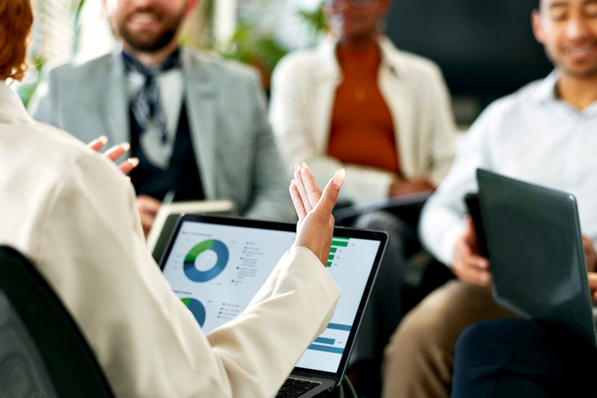 a diverse group of business professionals gathered around a laptop displaying data analytics charts and pie graphs, actively…