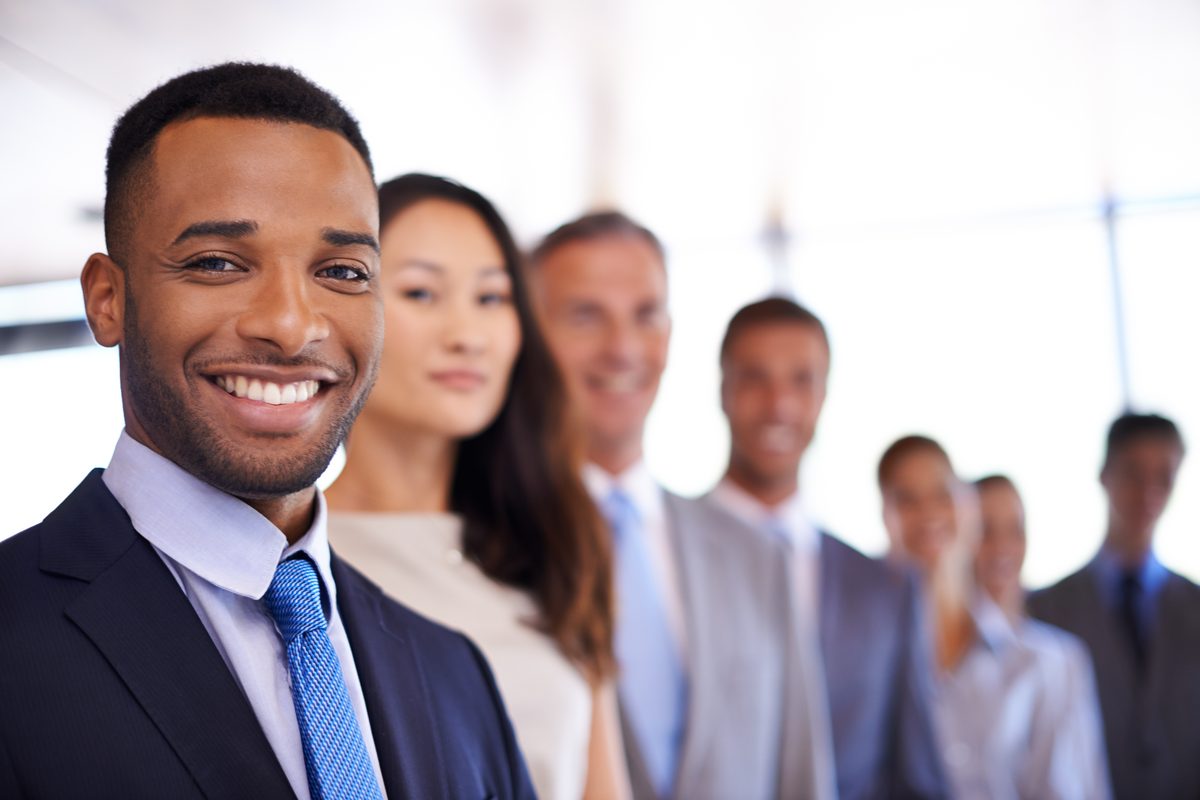 a diverse group of professional business people in formal attire standing together, with a smiling man in a dark suit and…