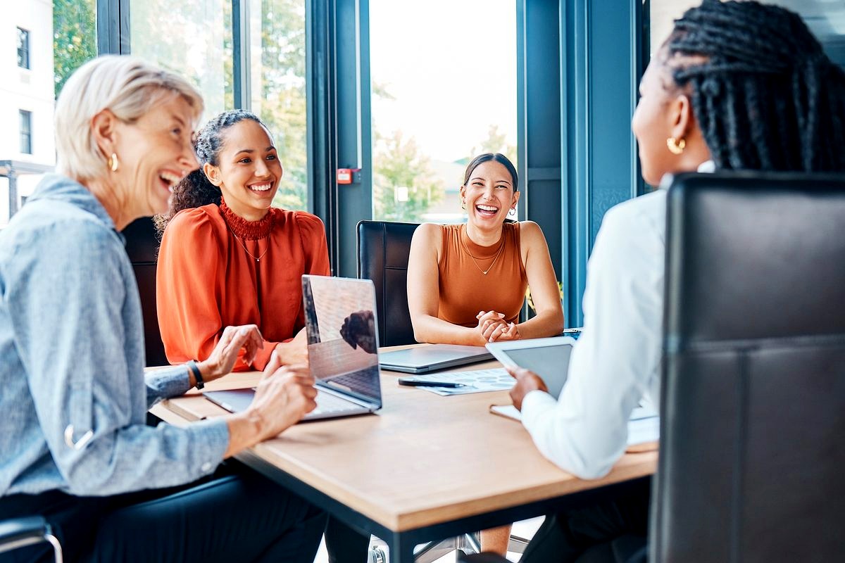 # Image Description

This image shows a diverse group of five women engaged in an animated, cheerful discussion around a…