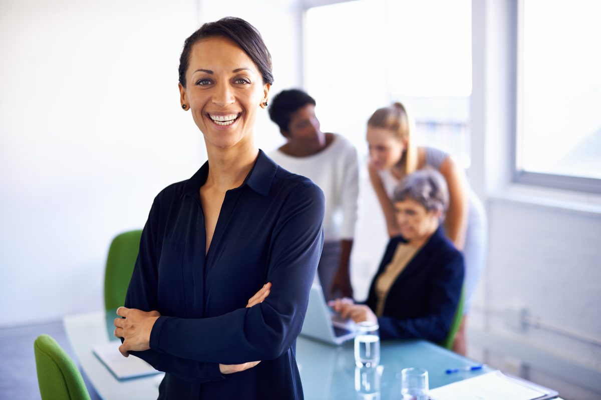 # Image Description

This photograph shows a confident, smiling businesswoman in professional attire standing with arms…