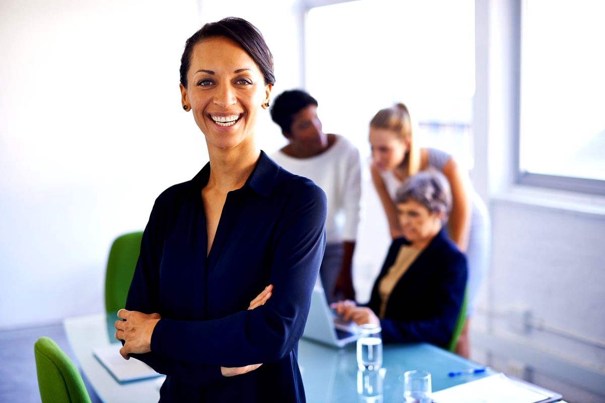 # Image Description

This photograph shows a confident, smiling businesswoman in professional attire standing with arms…