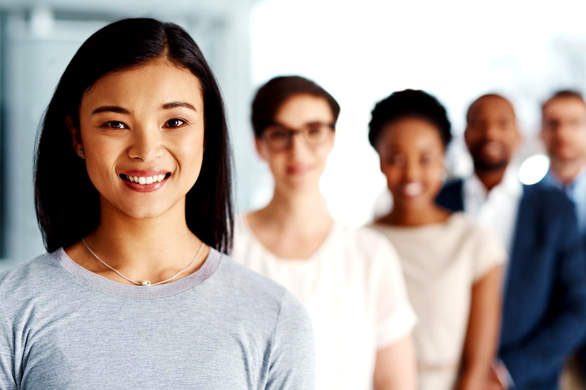 a smiling young Asian woman in sharp focus standing in front of a blurred group of diverse professionals in an office…