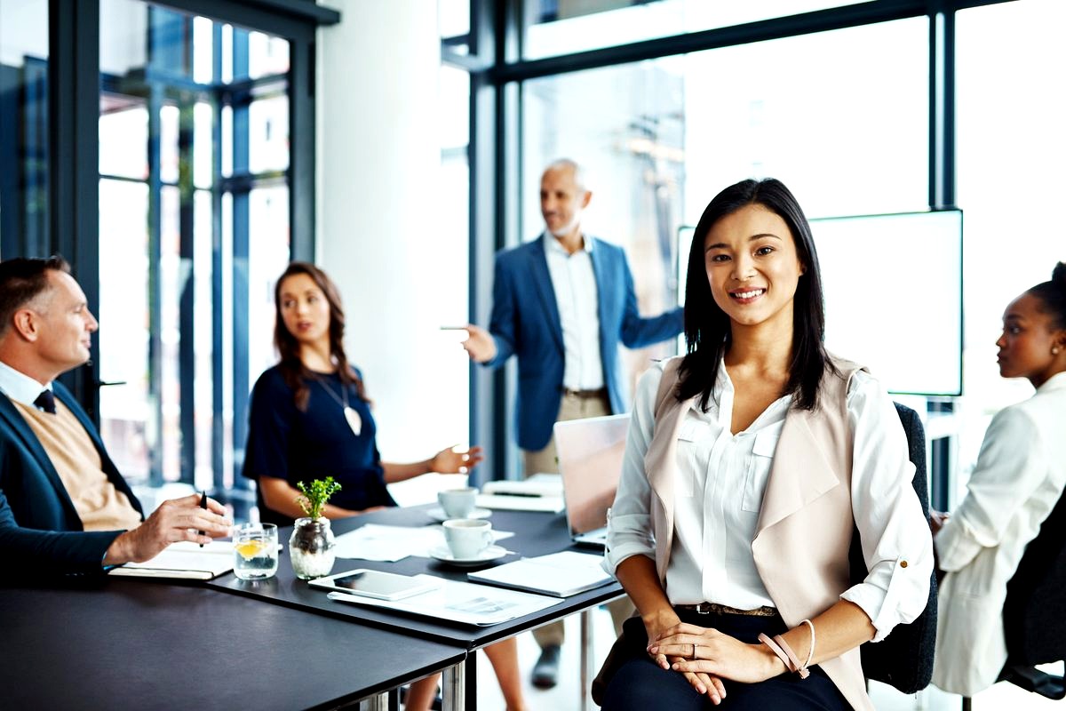 a confident young Asian woman in professional business attire smiling at the camera while standing in a modern conference…