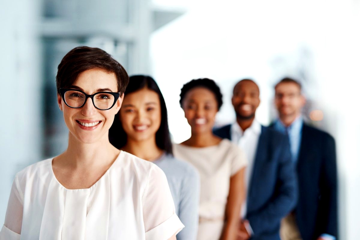 a diverse group of five smiling professionals standing in a line in a modern office setting, with the woman in front wearing…