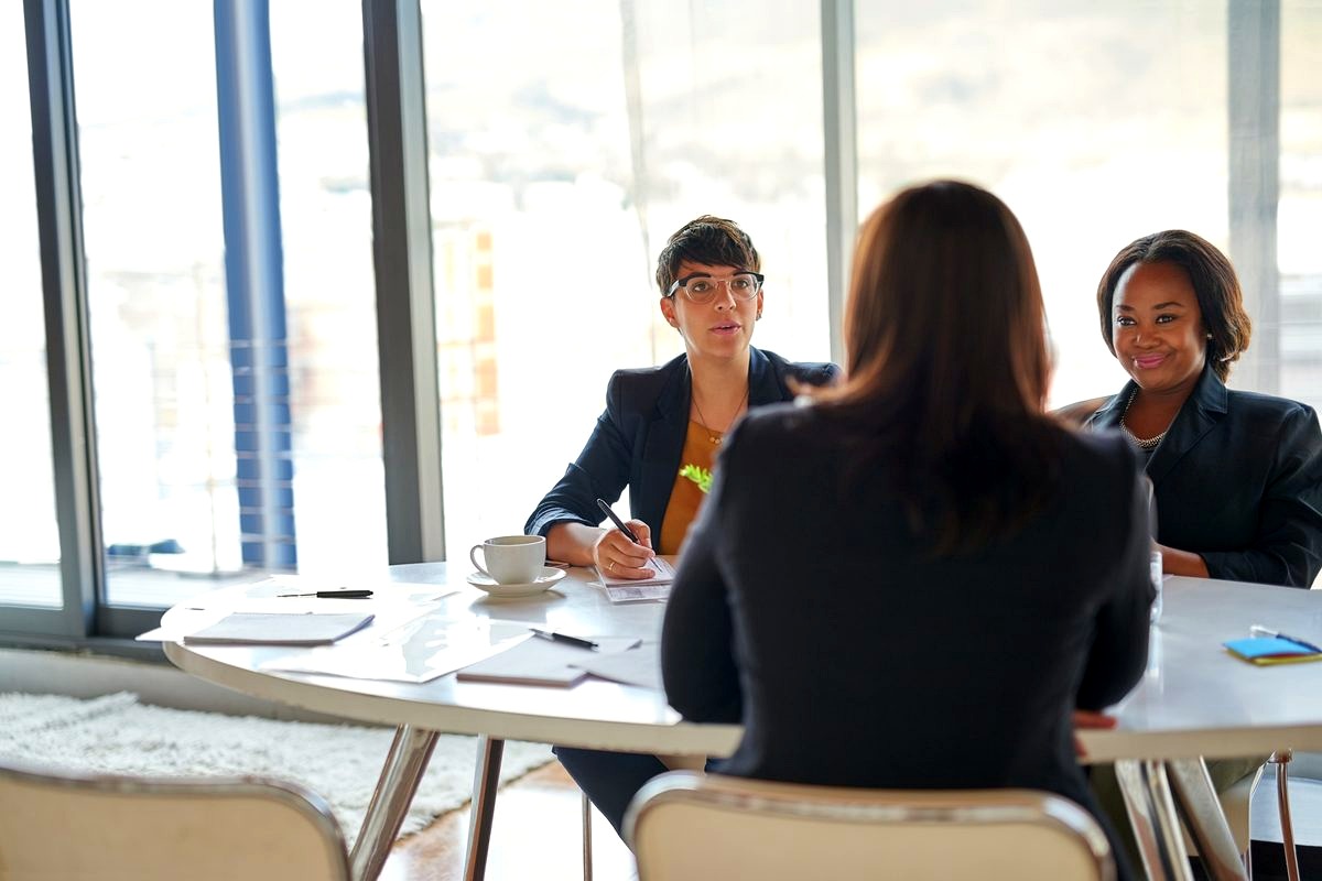 a professional job interview or panel discussion taking place in a modern high-rise office with floor-to-ceiling windows…