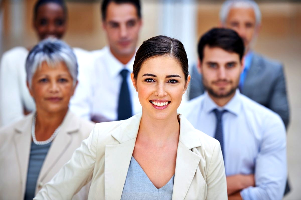 # Image Description
This professional photograph depicts a confident young woman in a cream blazer smiling at the camera…